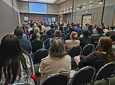 Large audience seated in a conference room listening to a presentation, with a speaker at the front and information displayed on a projection screen.
