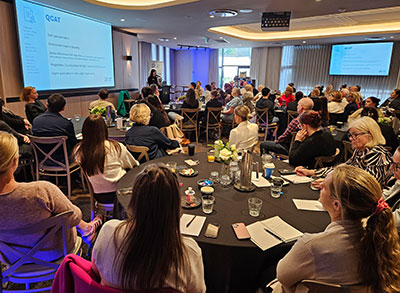 Continuing professional development attendees seated at round tables listen to a presentation, with slides displayed on large screens at the front of the room.