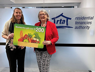 Two people stand in front of a Residential Tenancies Authority office sign, holding a large promotional sign for a $200 Woolworths gift card prize.