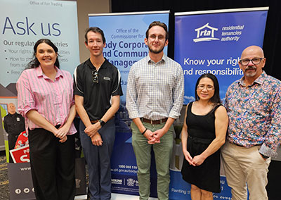 Five representatives stand in front of information banners from the Office of Fair Trading, the Office of the Commissioner for Body Corporate and Community Management, and the Residential Tenancies Authority at the collaborative information session.