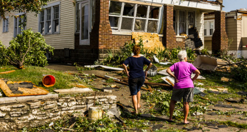 Two women standing on the front lawn of a house damaged by natural disaster.
