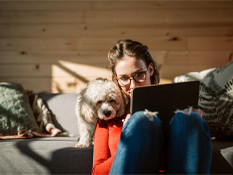 Young woman works on computer with dog leaning on her shoulder watching the screen.